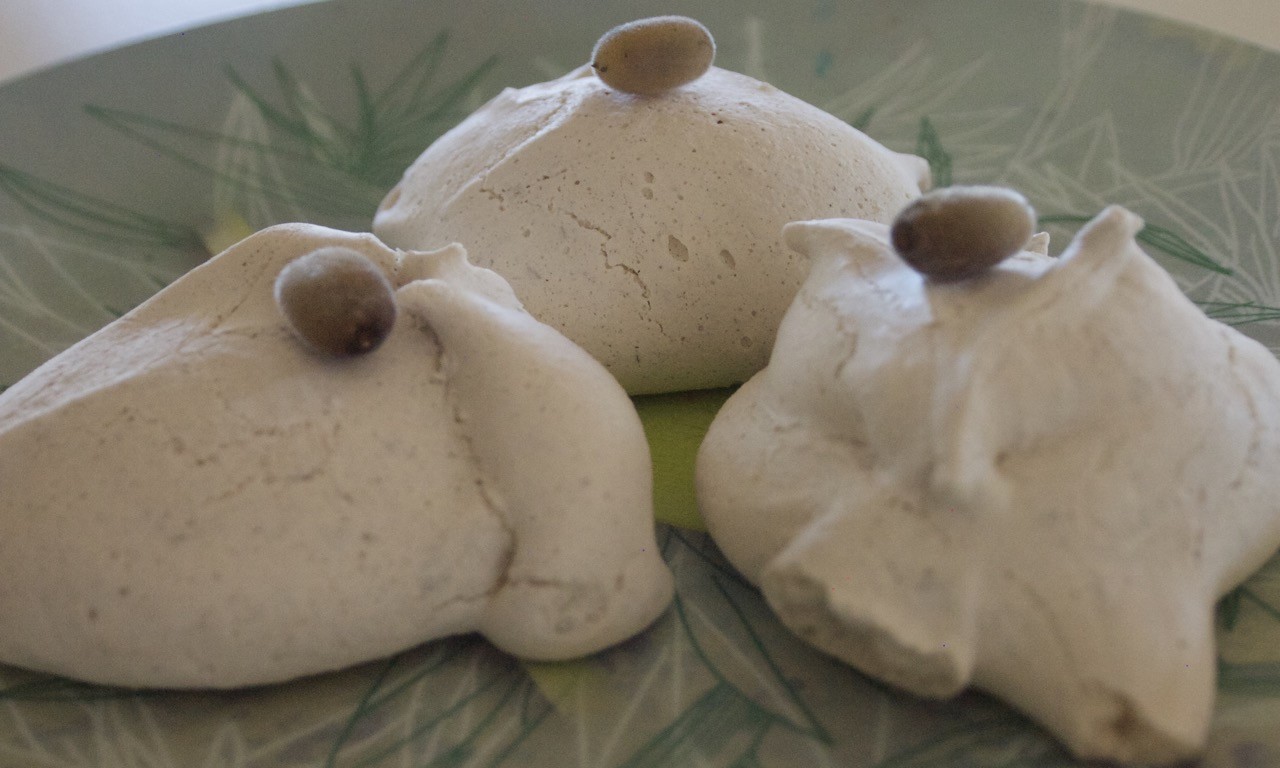Three white meringues sit on a pale green plate. They have a small berry balanced on top.