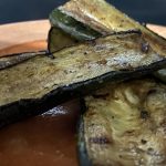 A close-up of zucchini sliced lengthwise. It is grilled, with slightly dark edges, and dark speckles on the slices, which is the remnants of the salty marinade. Three slices are sitting on an orange plate.