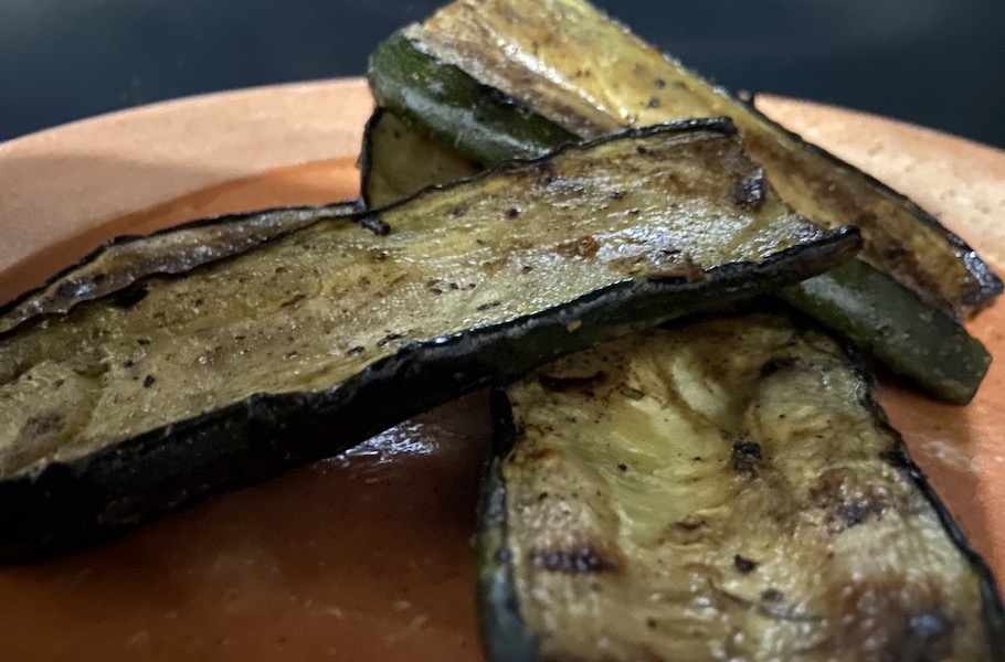 A close-up of zucchini sliced lengthwise. It is grilled, with slightly dark edges, and dark speckles on the slices, which is the remnants of the salty marinade. Three slices are sitting on an orange plate.