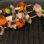 Five kebabs sit on a grilling top. They are strung with vegetables - whole cherry tomatoes, onion, capsicum, brussel sprouts, spring onions - haloumi, and finished with a folded leaf at the top. They are clearly cooked, with dark brown crispy bits on the veggies.