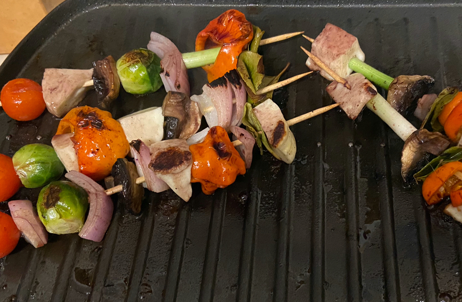 Five kebabs sit on a grilling top. They are strung with vegetables - whole cherry tomatoes, onion, capsicum, brussel sprouts, spring onions - haloumi, and finished with a folded leaf at the top. They are clearly cooked, with dark brown crispy bits on the veggies.