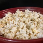 White popcorn is piled in a deep red bowl. There are dark specks of salt and herbs on some grains.